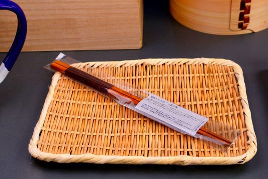 saibashi cooking chopsticks made of bamboo with urushi coating wrapped in plastic packaging atop a bamboo plate grey background