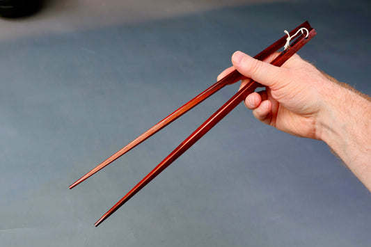 hand holding long square brownish tan hinoki wood cooking chopsticks urushi lacquered connected with white twine grey backdop