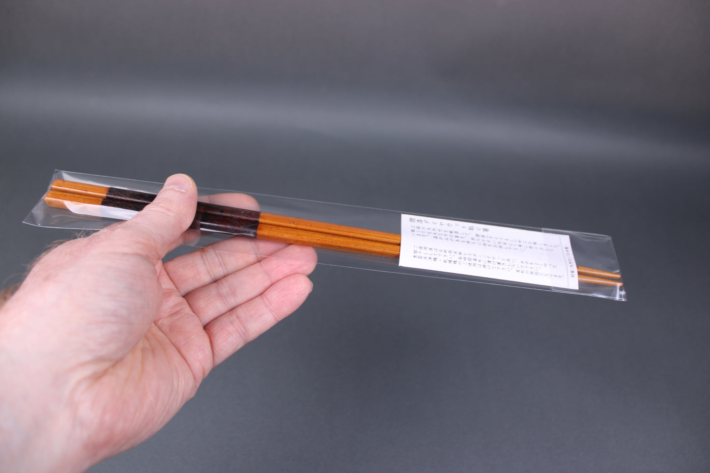 hand holding long square brownish tan bamboo cooking chopsticks urushi lacquered in clear packaging white product paper description in hiragana grey backdop
