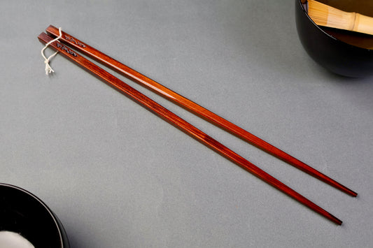 long square brownish tan hinoki wood cooking chopsticks urushi lacquered connected with white twine surrounded by japanese urushi bowls grey backdop