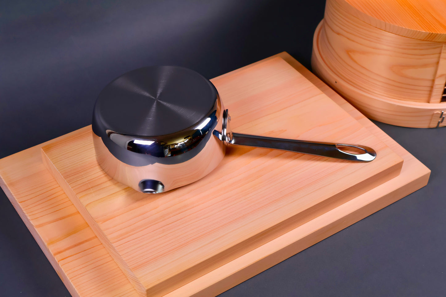 underside of yukihira saucepan with spouted edge on wood boards beside round wood steamer in grey background