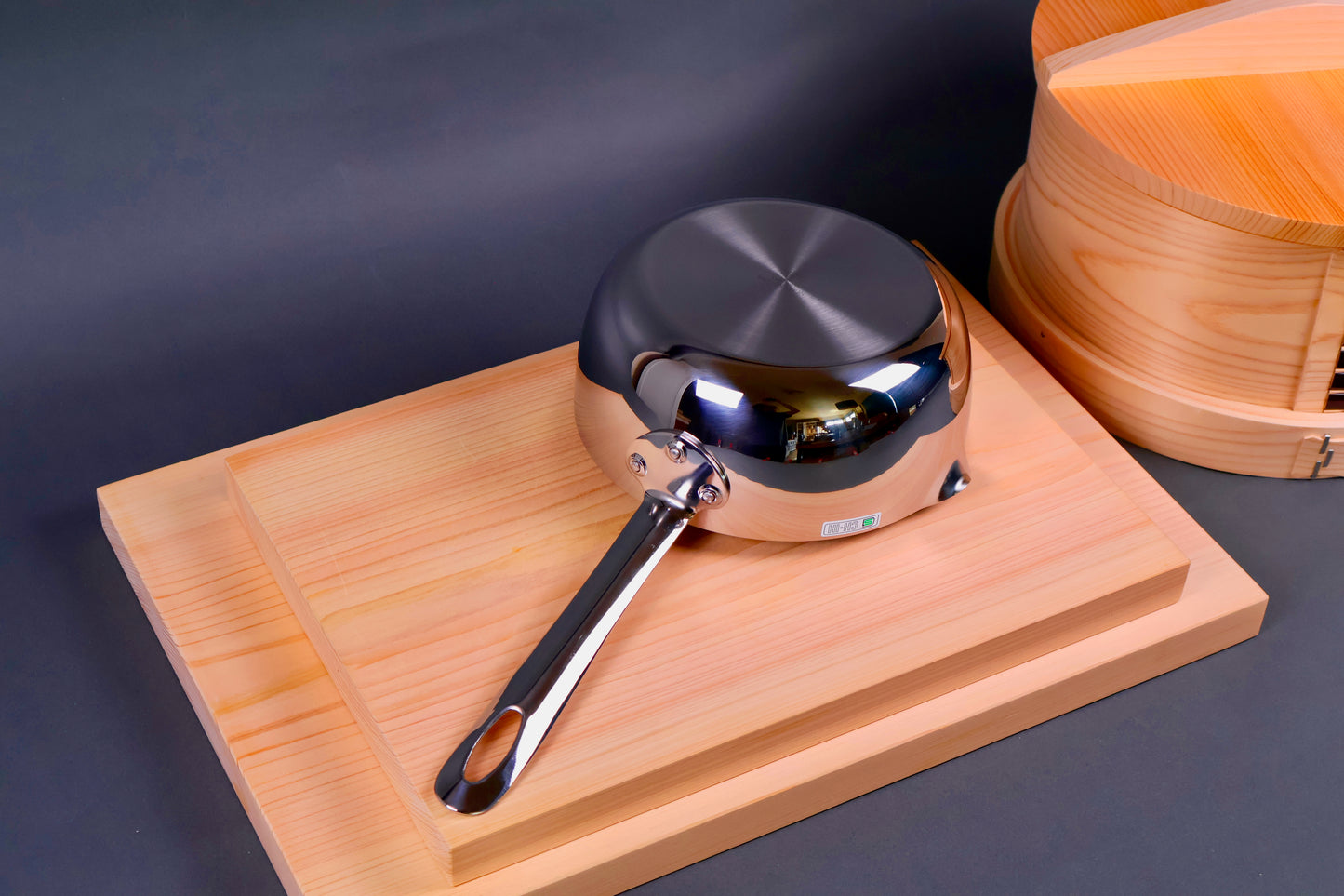 japanese steel saucepan underside showing elegant handle attached with rivets on wood boards next to round wood steamer grey backdrop