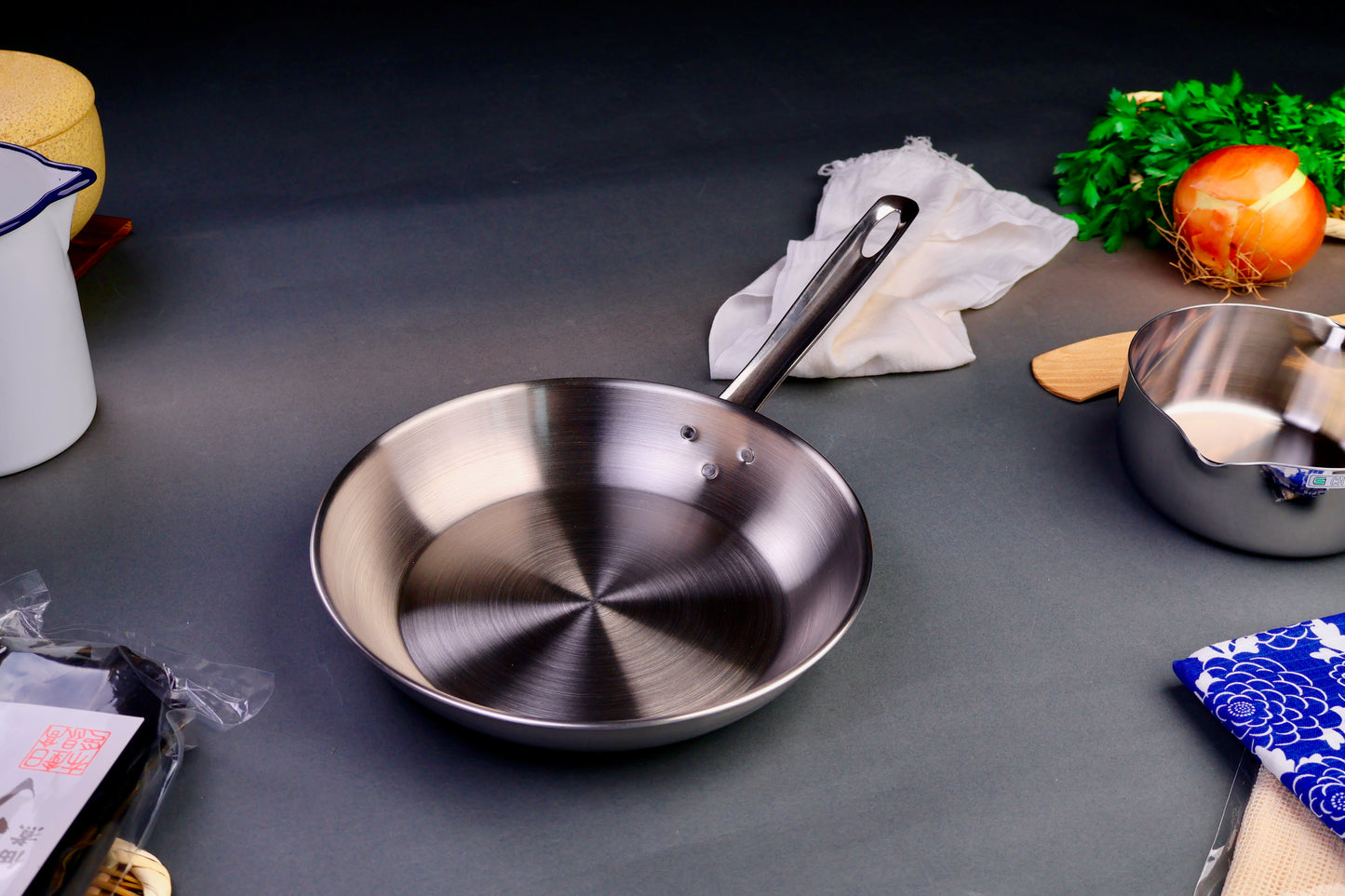 high quality stainless steel saute pan with long elegant handle surrounded by Japanese kitchenware with light grey background