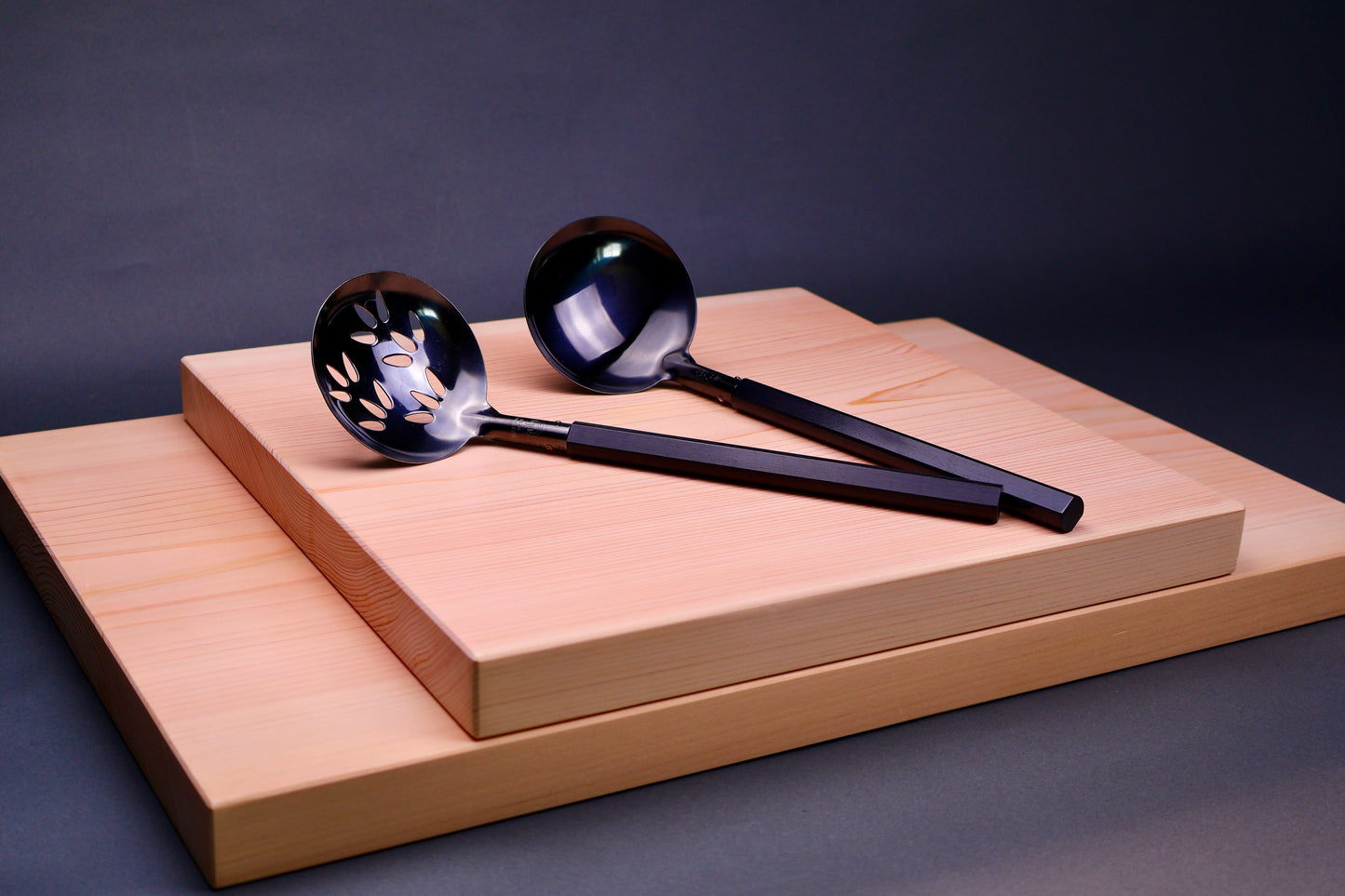 oxidized steel ladles one slotted one solid with wood handles sit atop two hinoki cutting boards with dark grey background