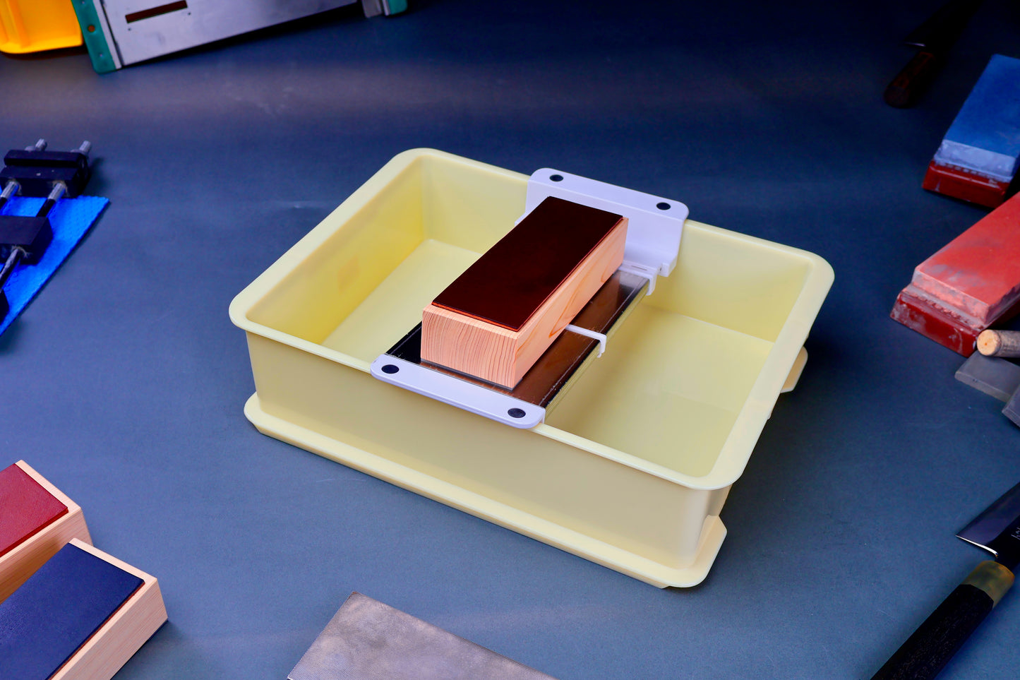 short yellow plastic container with suehiro multi sliding rack and hinoki leather knife strop surrounded by knives and accessories on blue grey surface