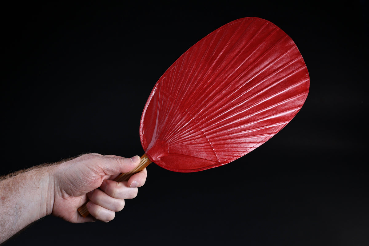 Shibu grilling fan red held in hand against black background, showing full oval fan face and bamboo handle