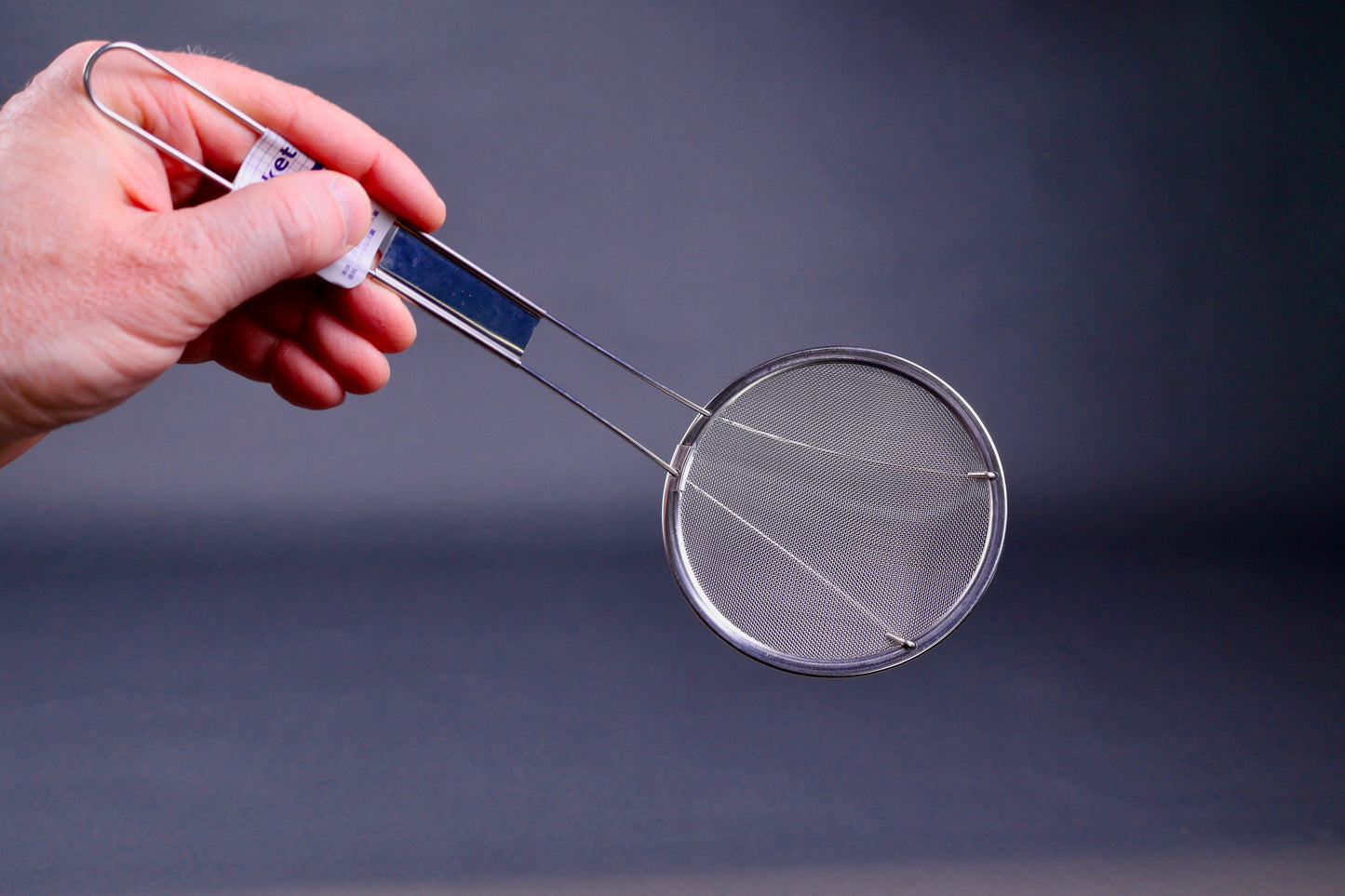 hand holding long handled stainless steel fine mesh skimmer showing thin wire in grey background
