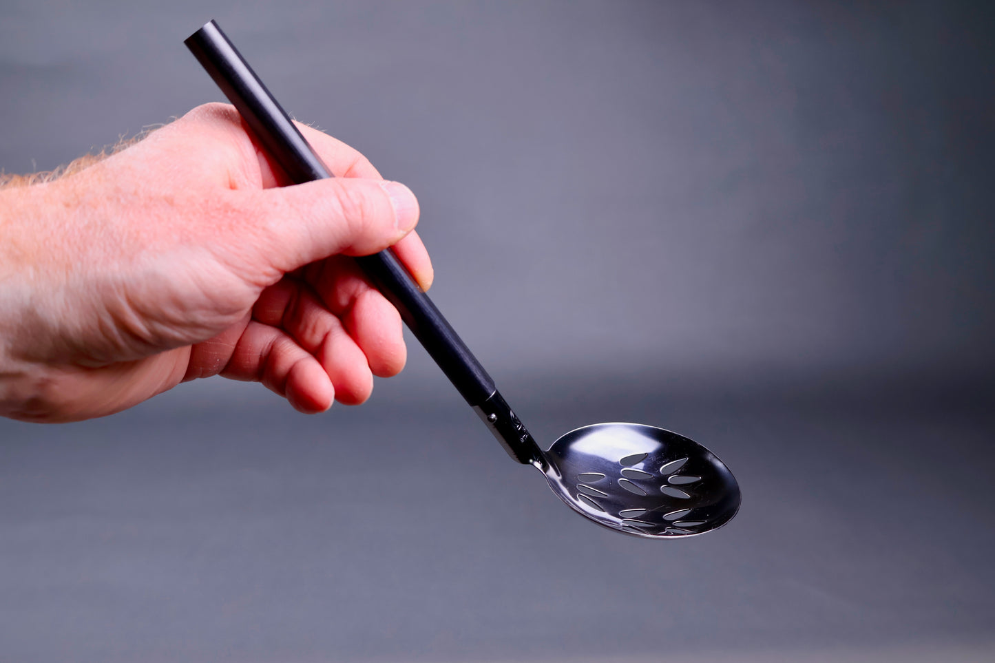 hand holdng a serving ladle perforated with slots in the metal grey background