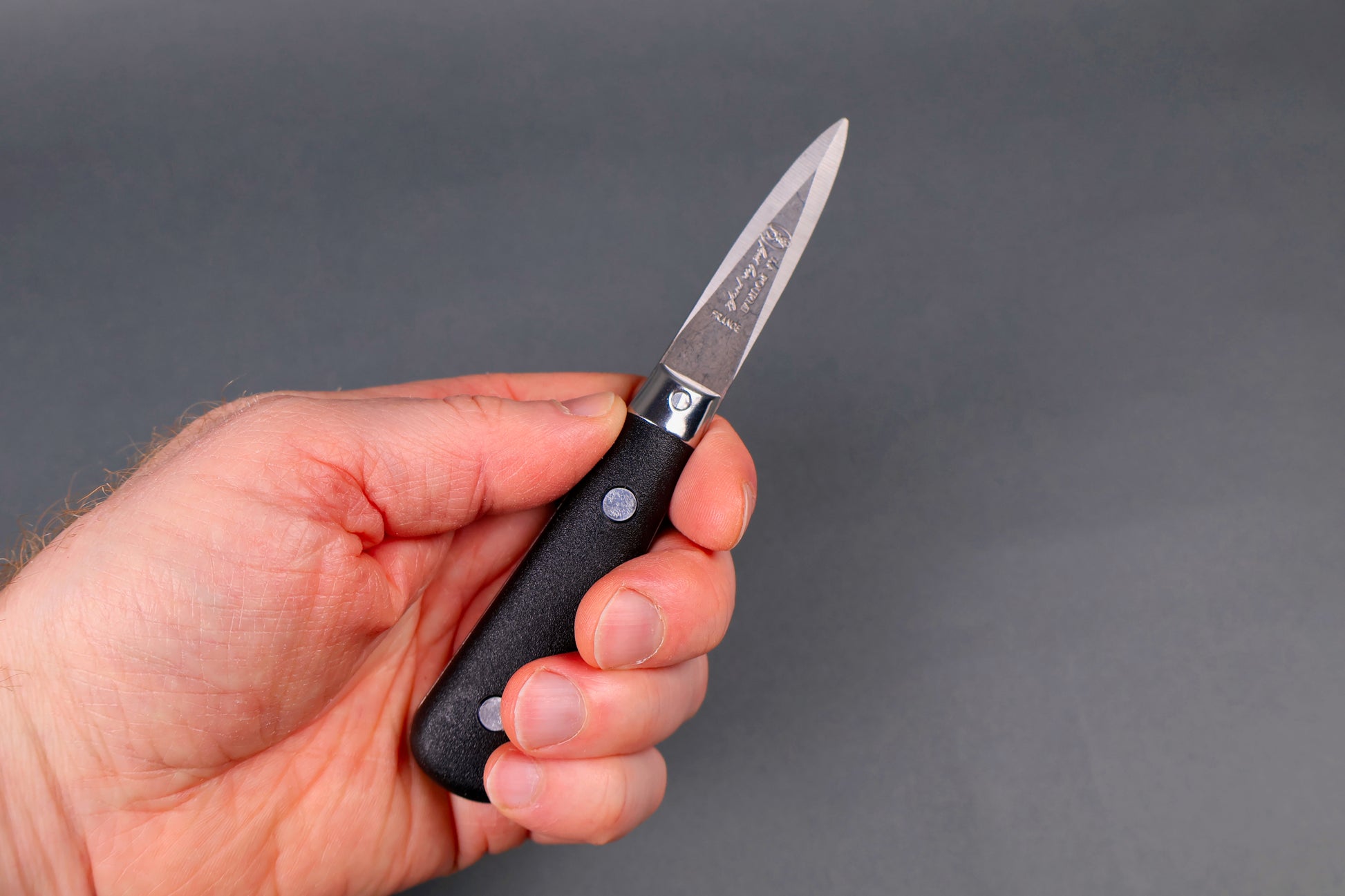 hand holding small black handled stainless steel oyster knife etched in French writing with beveled edges in grey background