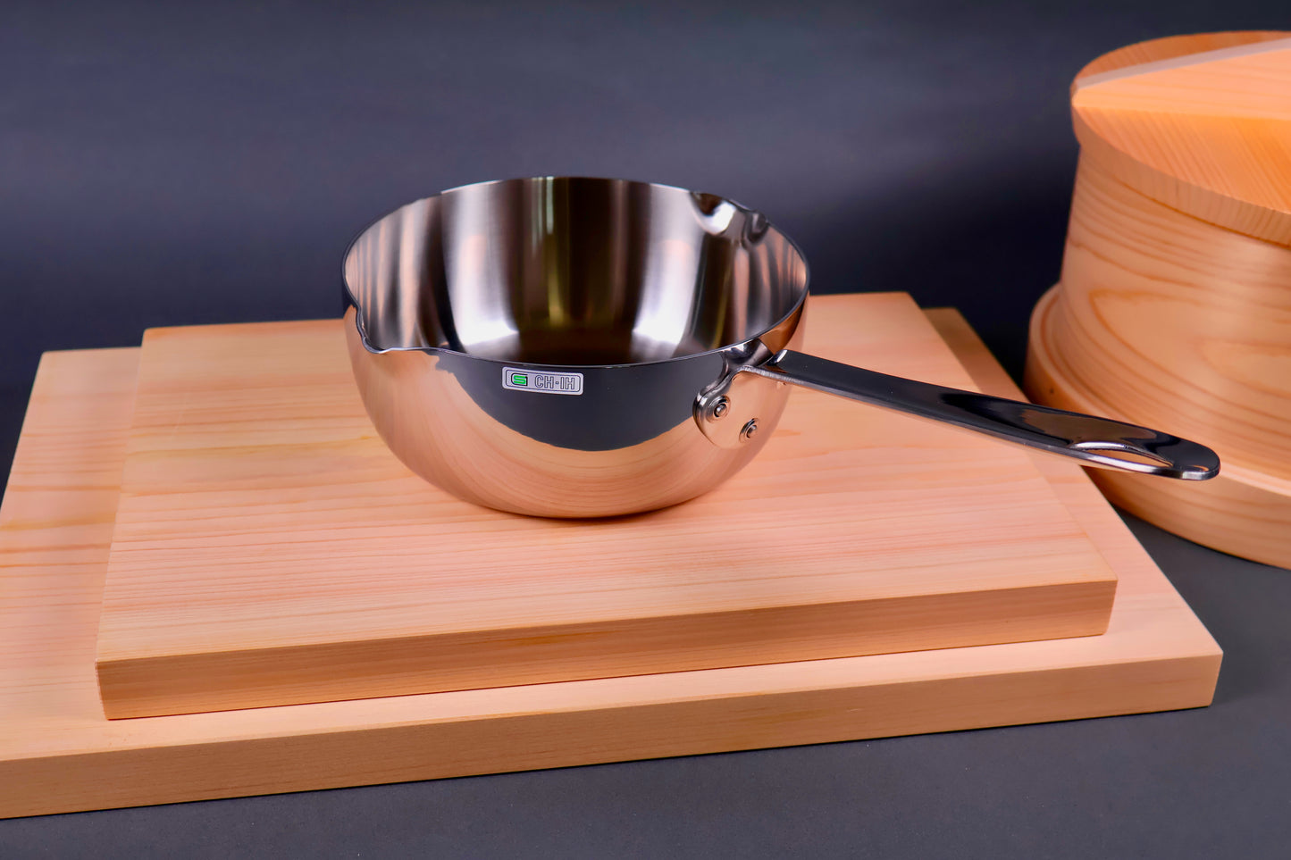shiny silver metal pan with long handle on wood boards beside round wood steamer in grey background