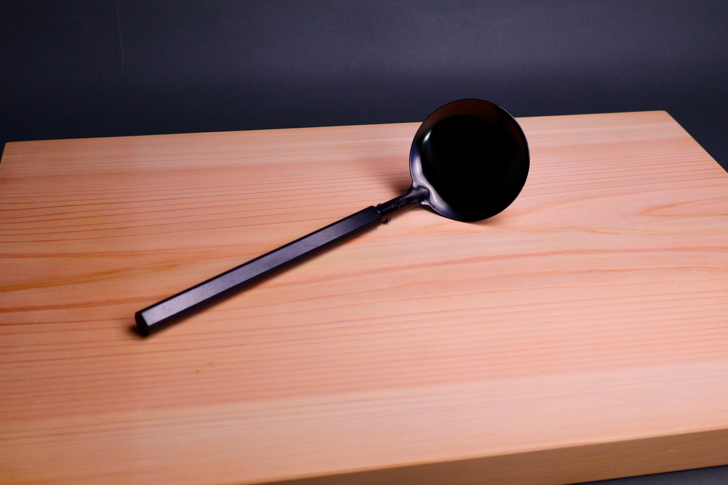 Black ladle with pakka wood handle on a wooden cutting board with a dark background greyish