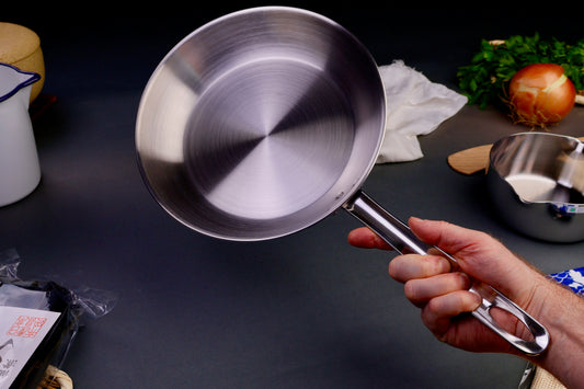hand holding long handled saute pan surrounded by Japanese kitchenware with light grey background