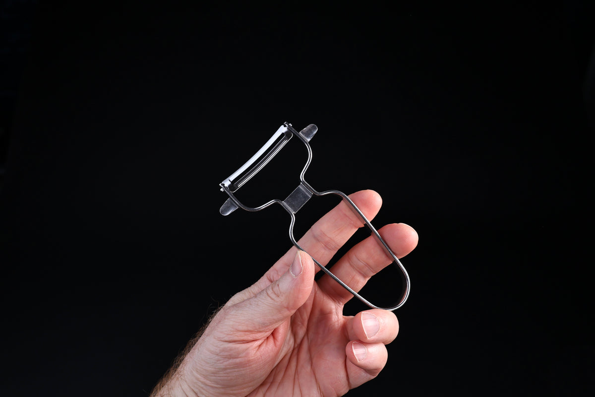 Stainless steel vegetable peeler — blade face, floating blade mechanism, and wire handle from reverse angle in hand