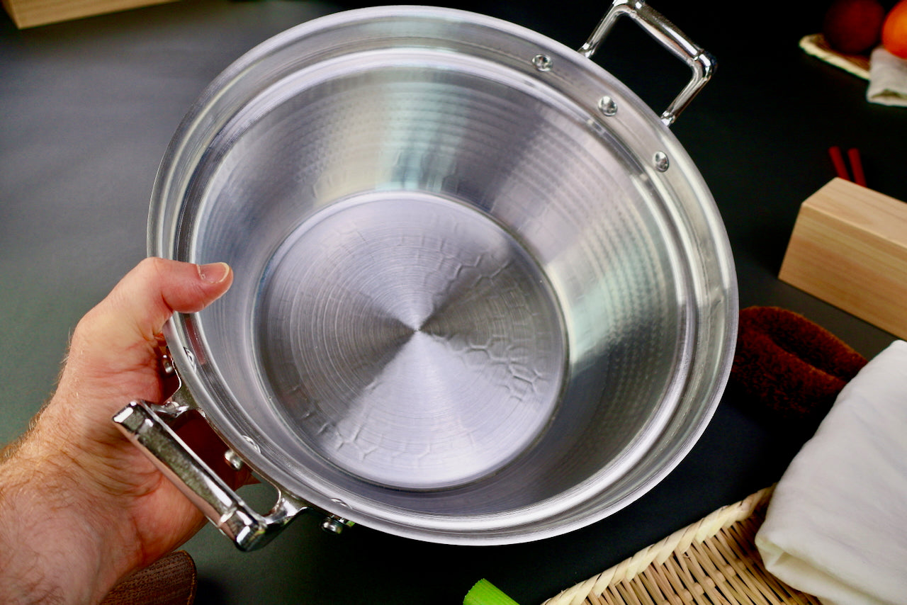 closeup hand holding two handled stepped aluminum silvery pot with shiny hammered surface surrounded by japanese kitchenware on a dark grey backdrop