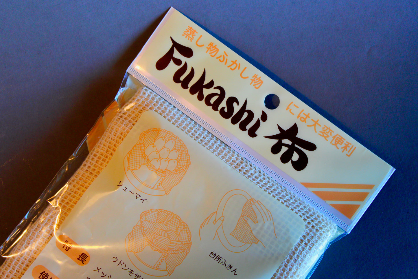 detail of long rectangular product plastic wrapped steam cloth with printed name in hiragana animated steamer with tan and yellow stripes on grey backdrop