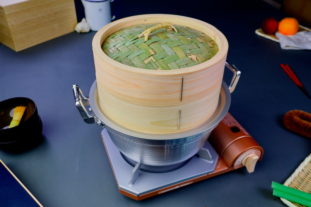 thick wood round bamboo steamer set woven top with loop natural wood tone with printed hiragana sits atop double handled metal hammered pot atop portable butane stoves gray background