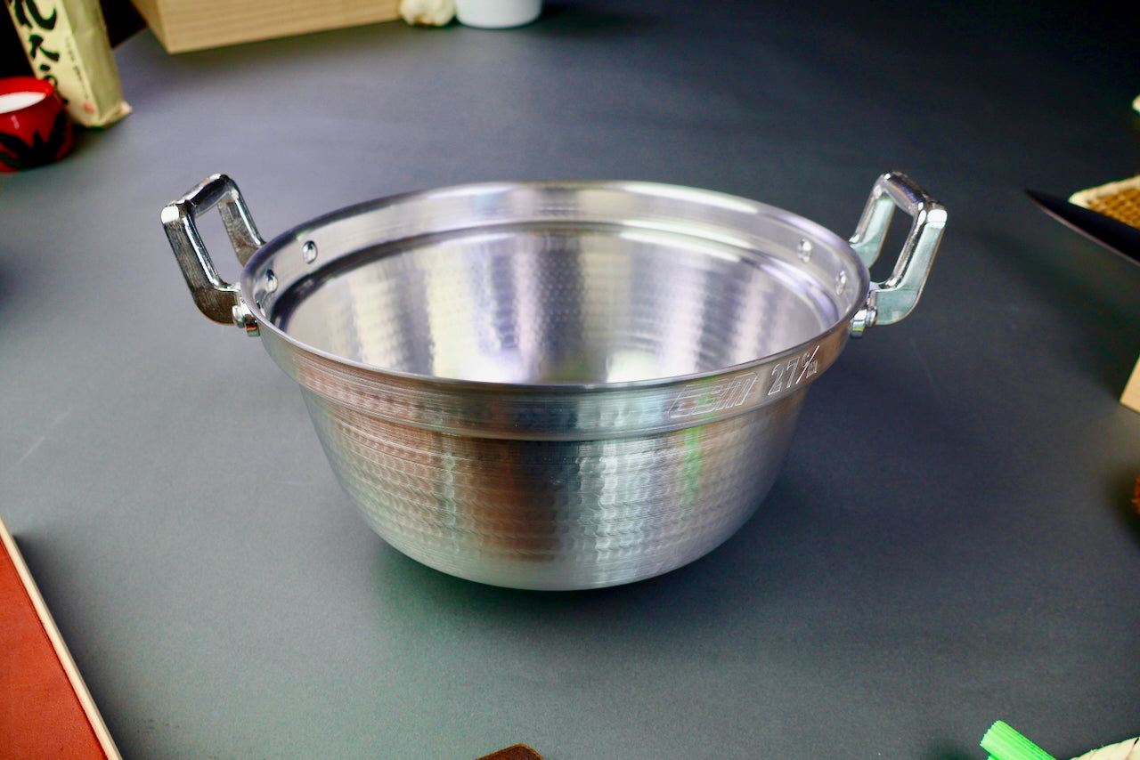 two handled stepped aluminum silvery pot with shiny hammered surface surrounded by japanese kitchenware on a dark grey backdrop