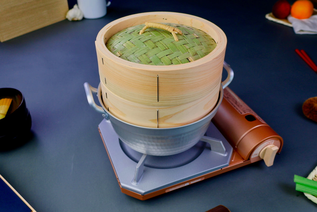 bamboo hinoki wood round steamer kit featuring thick round lid thick round base sitting neatly inside hammered pot on top of portable stove in bluish grey backdrop