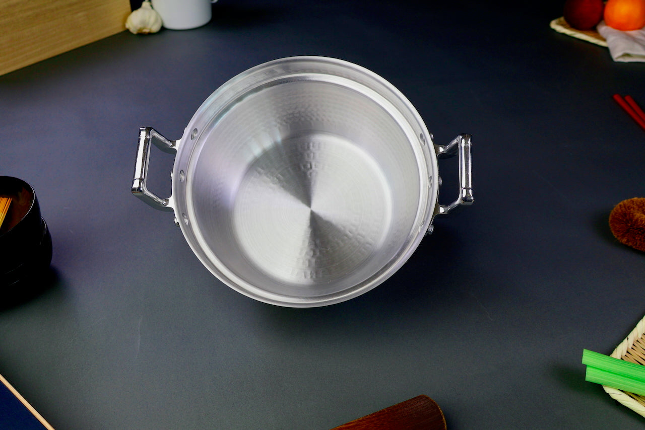 interior of two handled stepped aluminum silvery pot with shiny hammered surface surrounded by japanese kitchenware on a dark grey backdrop