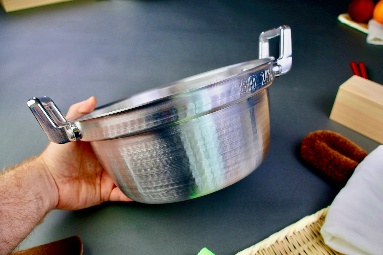 close up of hand holding two handled stepped aluminum silvery pot with shiny hammered surface dark grey backdrop
