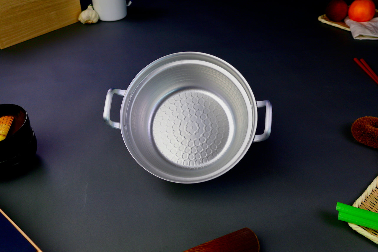 interior shot two handled stepped aluminum silvery pot with shiny hammered surface surrounded by japanese kitchenware on a dark grey backdrop