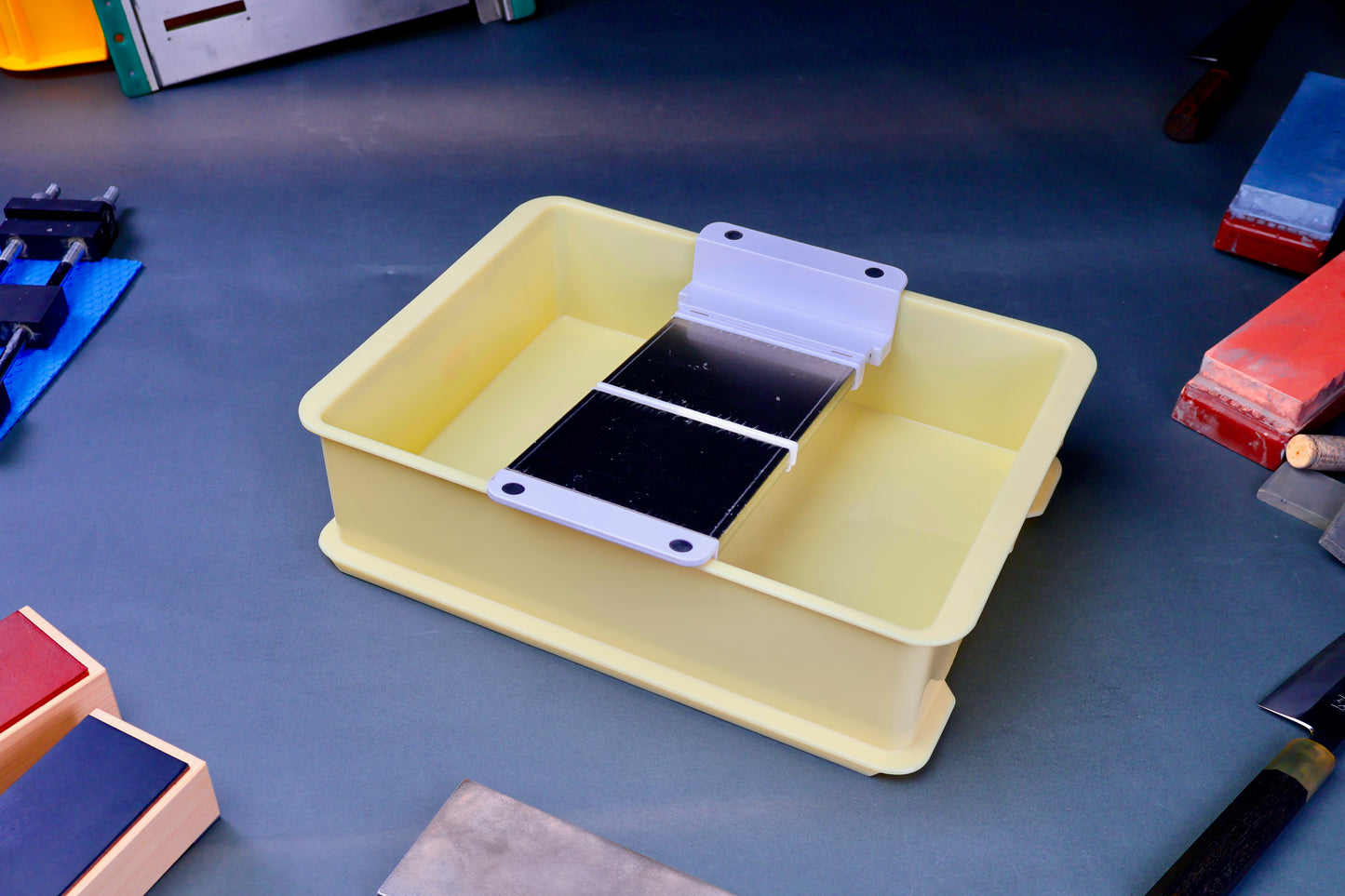 yellow plastic rectangular container holding suehiro sharpening accessory for whetstone surrounded by knife accessories on a grey blue surface