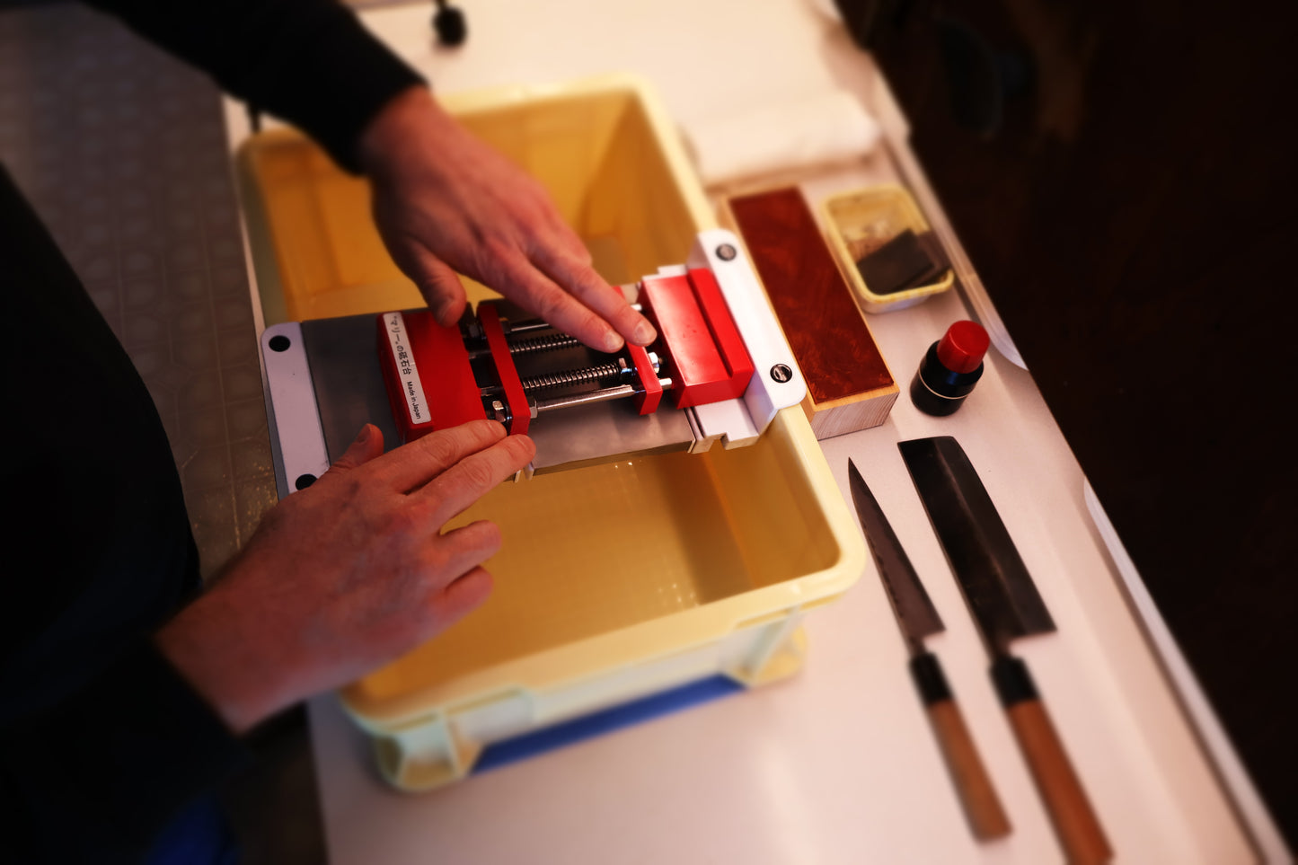kitchen counter with tall yellow container holding suehiro sliding rack with white edges atop sits marie whetstone holder red and two hands moving the product in place surrounded by knives