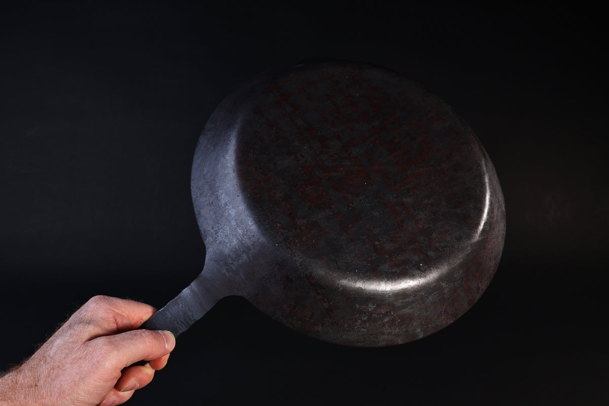 Turk hand-forged iron frying pan held by the handle showing the hammered underside against a dark background