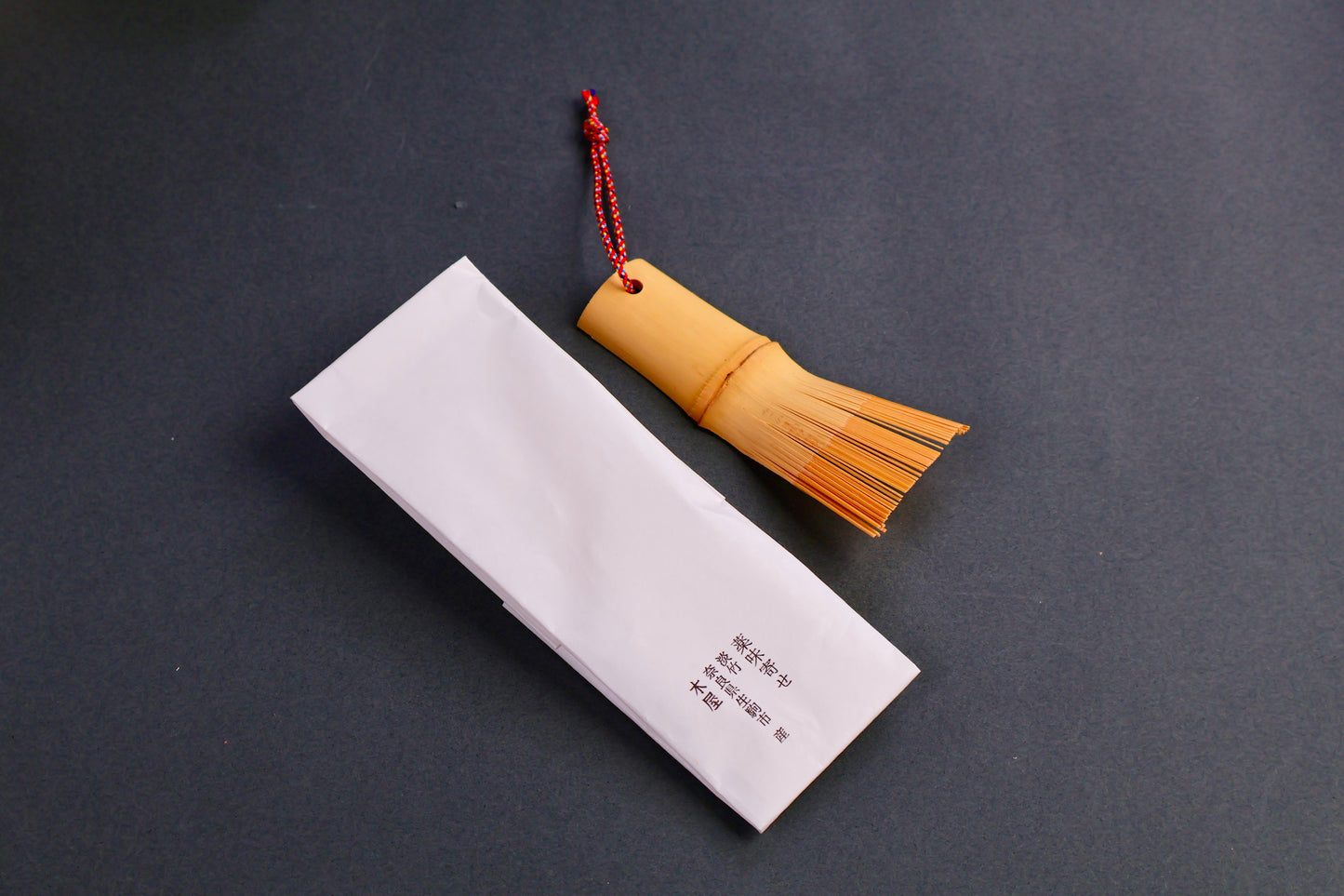 short rectangular bamboo brush with thin bristles red silk loop beside white rectangular paper wrapper with black kanji print on dark grey backdrop