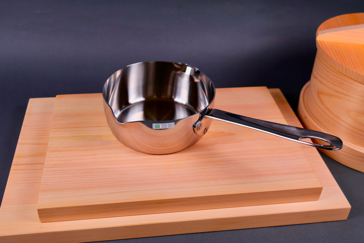 top down shot of long handled wide body beaked saucepan on top of hinoki wood cutting boards beside seiro steamer grey background