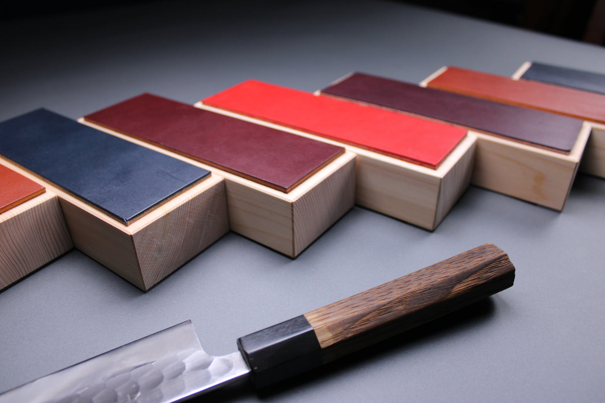 handmade leather knife strop, crafted from the highest grade english bridle leather with a hinoki wood base imported from nara prefecture made in a variety of colors displayed next to a japanese knife with grey background 