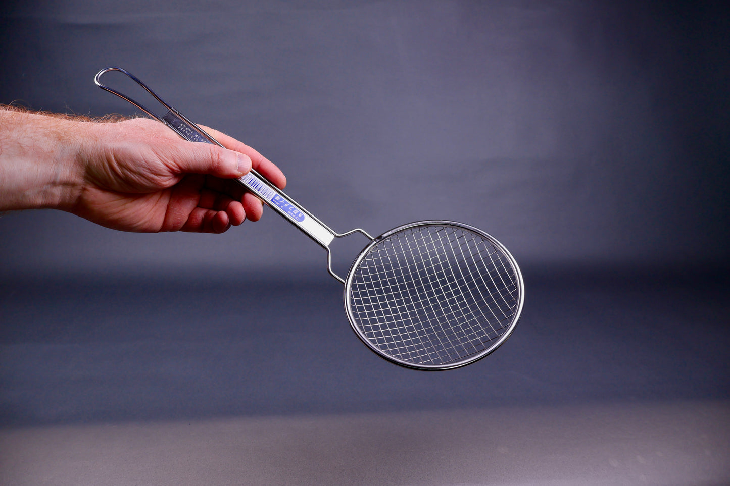 Hand holding a stainless steel coarse strainer with long sturdy handle against a dark background
