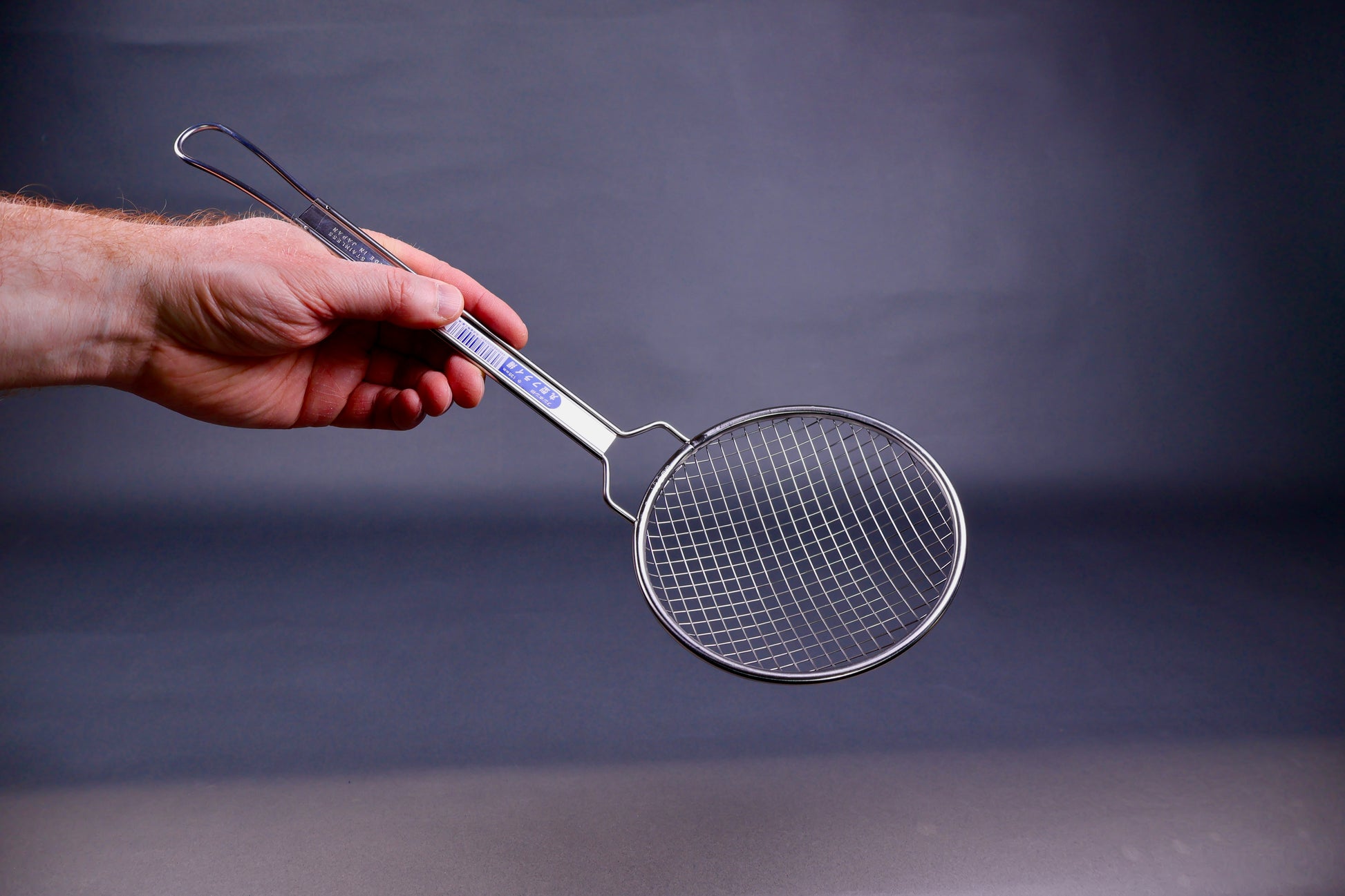 Hand holding a stainless steel coarse strainer with long sturdy handle against a dark background