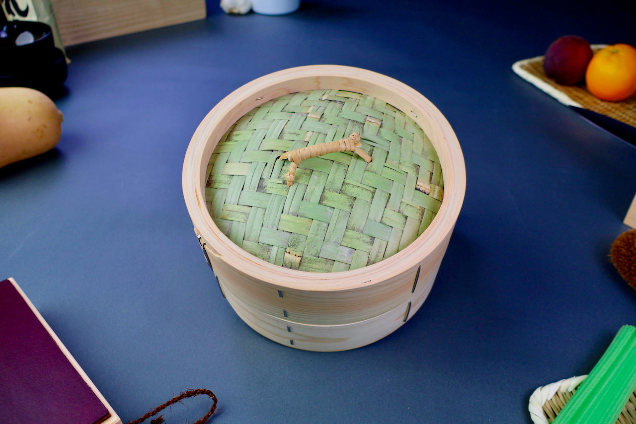 top down shot of thick round hinoki wood steamer surrounded by colorful kitchen ware fruit vegetables leather strop bluish backdrop