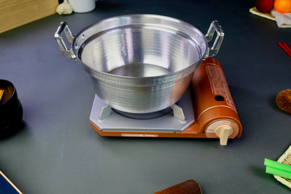 two handled stepped aluminum silvery pot with shiny hammered surface sits atop portable silver tan butane stove surrounding kitchenware dark grey backdrop 