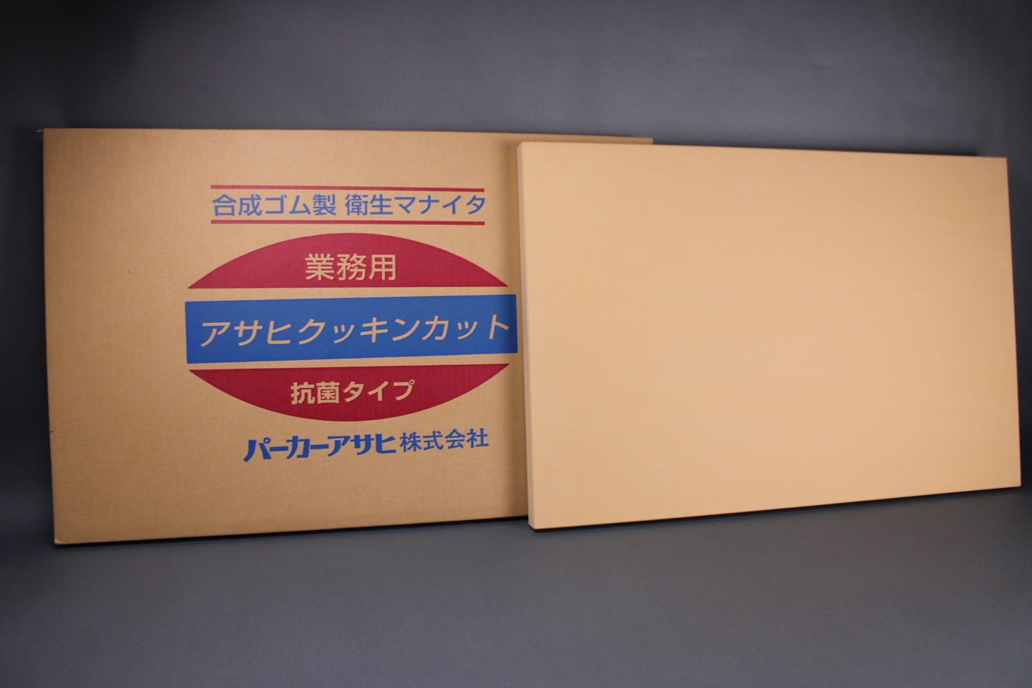 frontal shot of asahi rubber cutting board and packaging box with japanese writing showing the pleasant yellow shaded surface and large rectangular shape with grey background