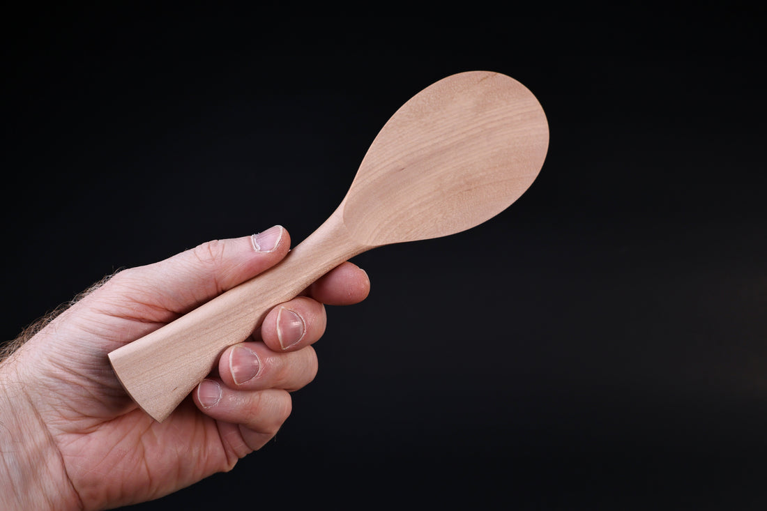 Azmaya handmade maple rice paddle held by the handle, paddle end facing up against a dark background
