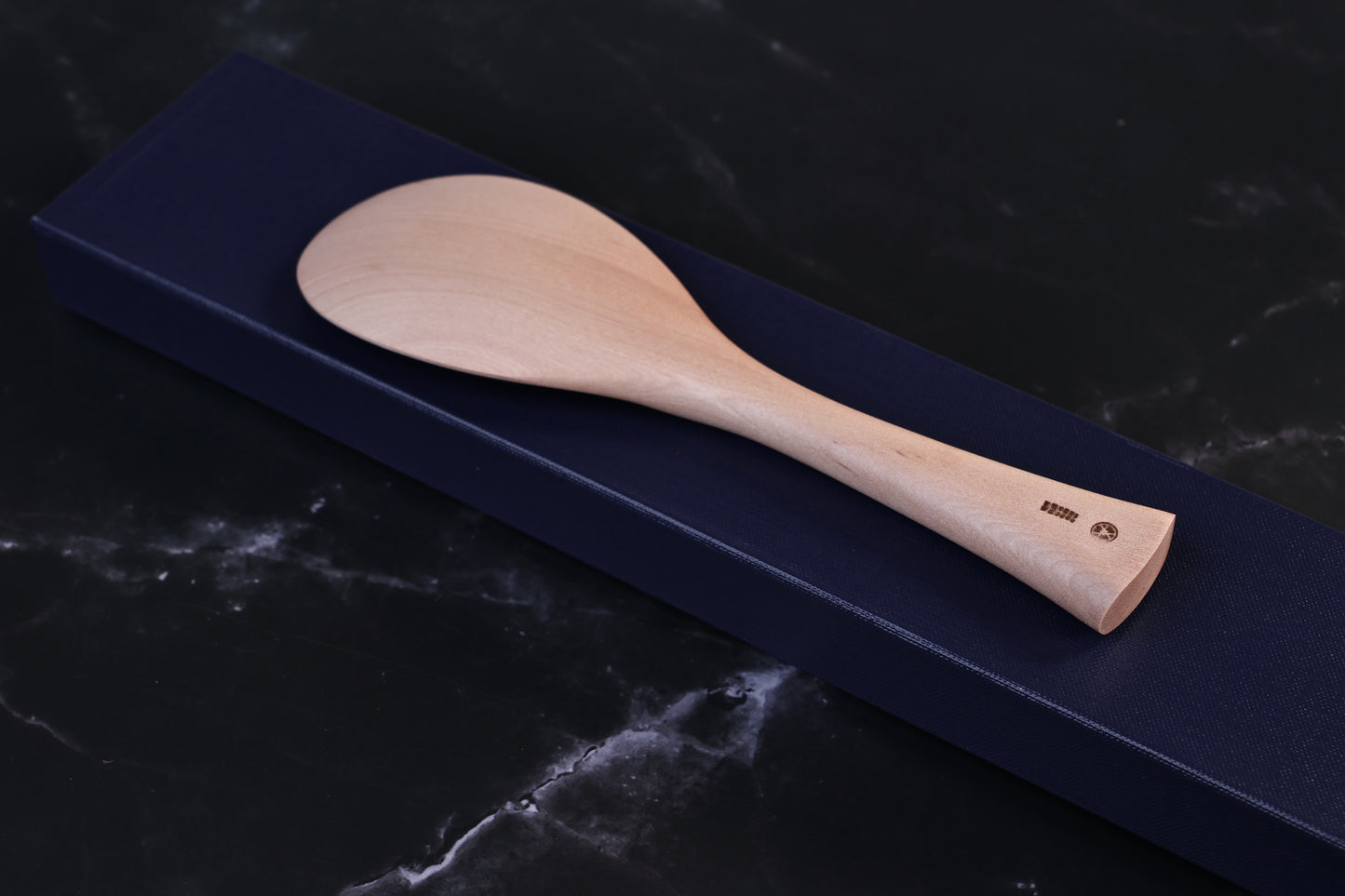 Azmaya handmade maple rice paddle laid across its navy gift box, shown at an angle with maker's marks visible