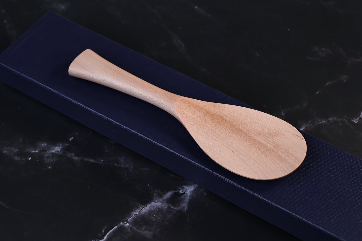Azmaya handmade maple rice paddle resting on its navy gift box on a dark marble surface