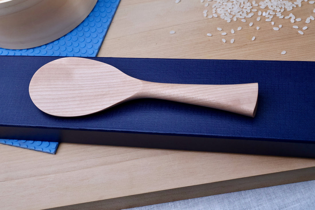Azmaya handmade wooden rice paddle on blue cutting board with scattered rice grains