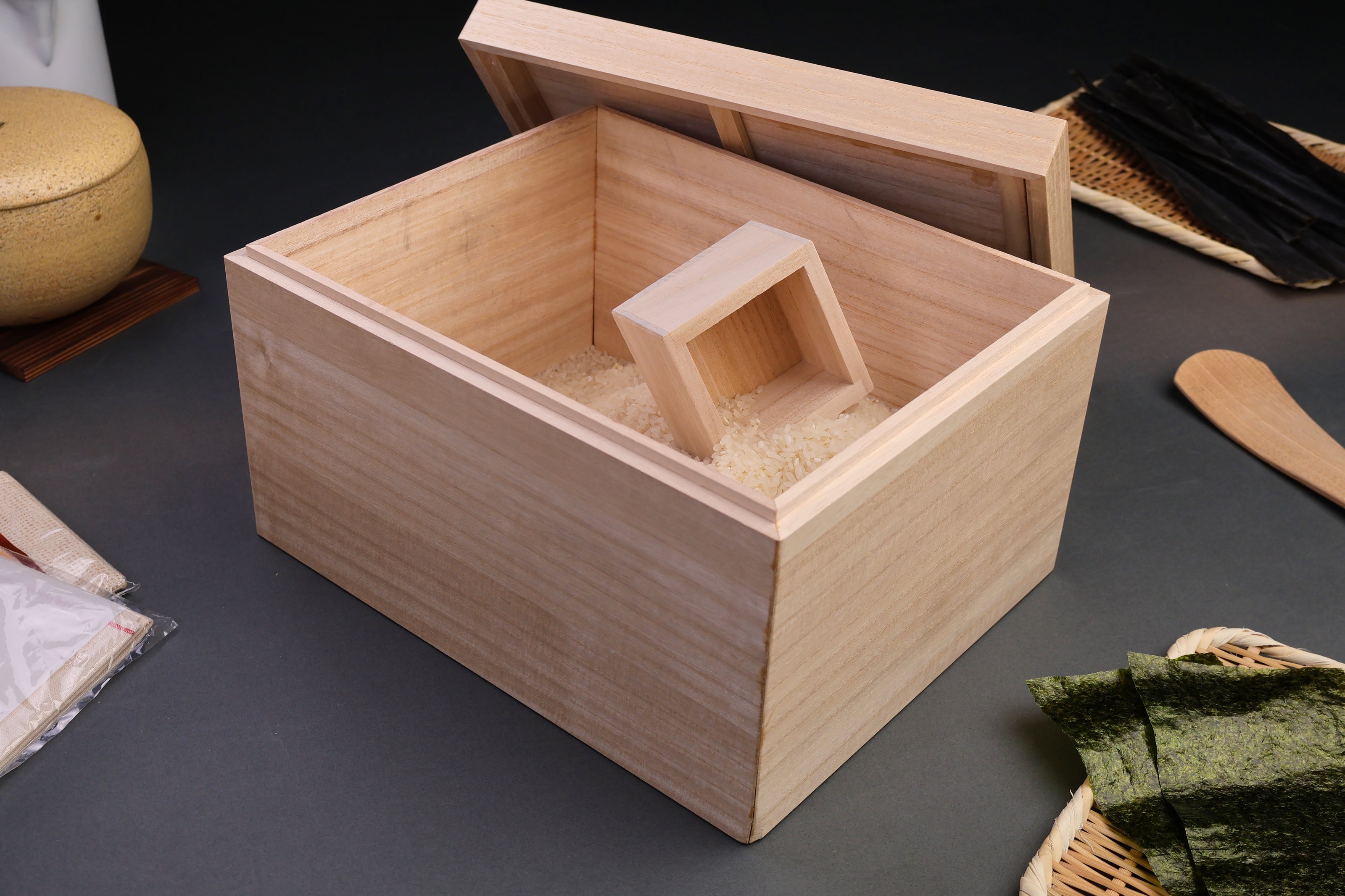 tall rectangular paulownia wood rice box sliding door on top removable with rice scoop surrounded by japanese kitchenware dark grey background