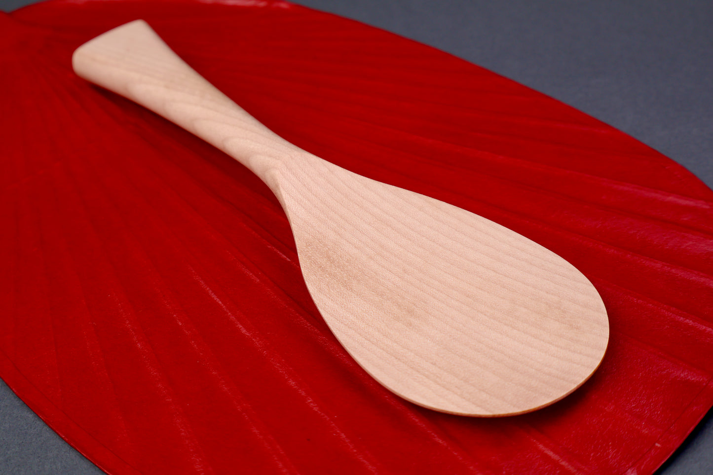 hand carved wooden rice paddle light golden brown yellowish tone of paddle atop deep red background surface