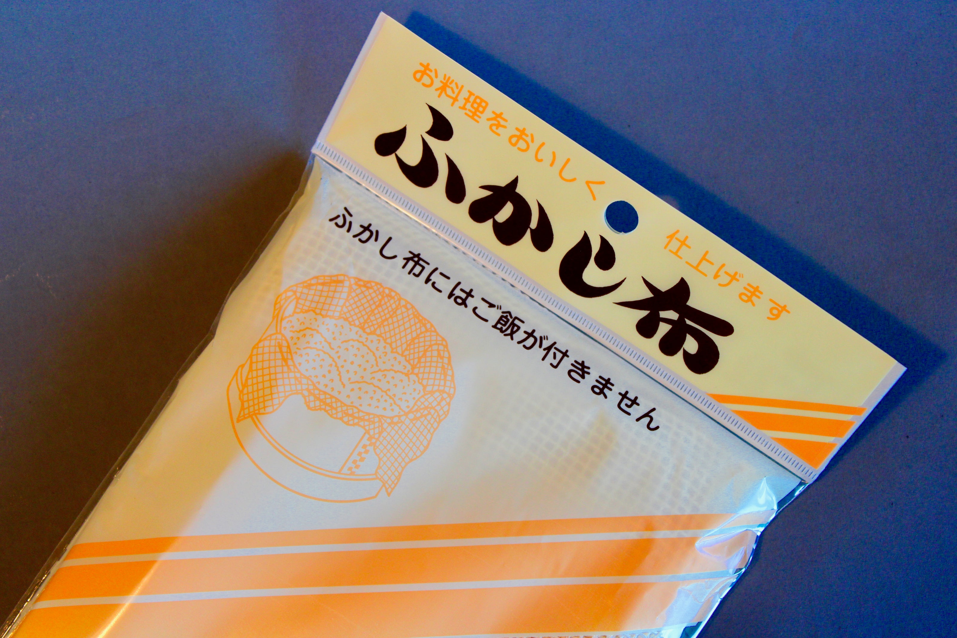 close up of long rectangular product plastic wrapped steam cloth with printed name in hiragana animated steamer with tan and yellow stripes on grey backdrop