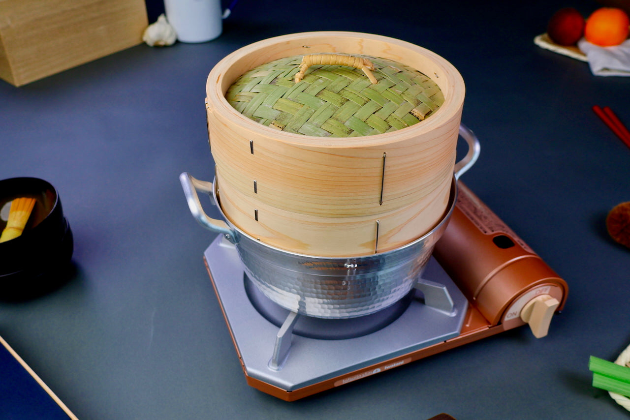 bamboo hinoki wood round steamer kit featuring thick round lid thick round base sitting neatly inside hammered pot on top of portable stove in bluish grey backdrop