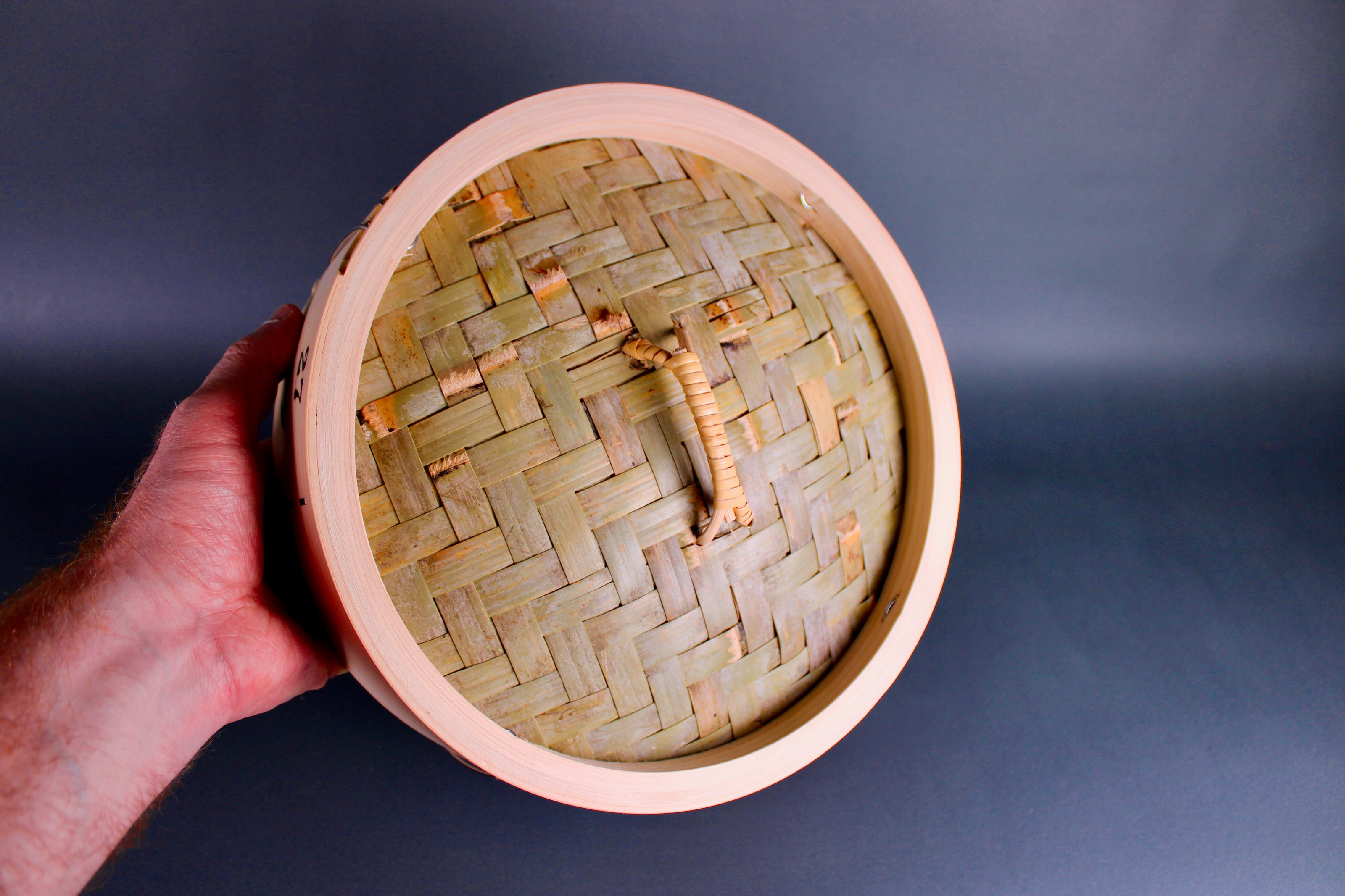 hand holding bamboo steamer lid woven bamboo with looped handle surrounded by thick hinoki wood exterior on gray surface