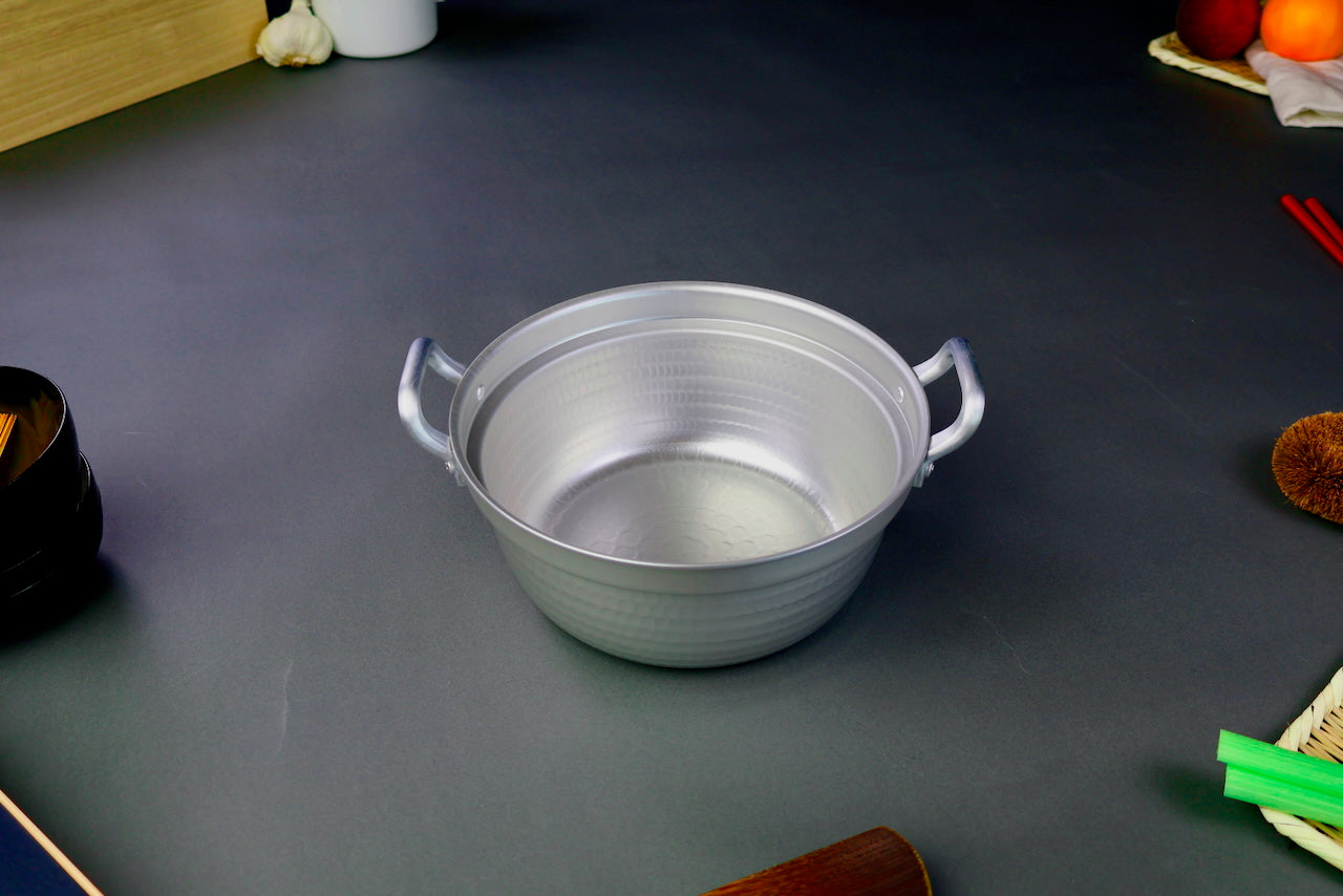 two handled stepped aluminum silvery pot with shiny hammered surface surrounded by japanese kitchenware on a dark grey backdrop 
