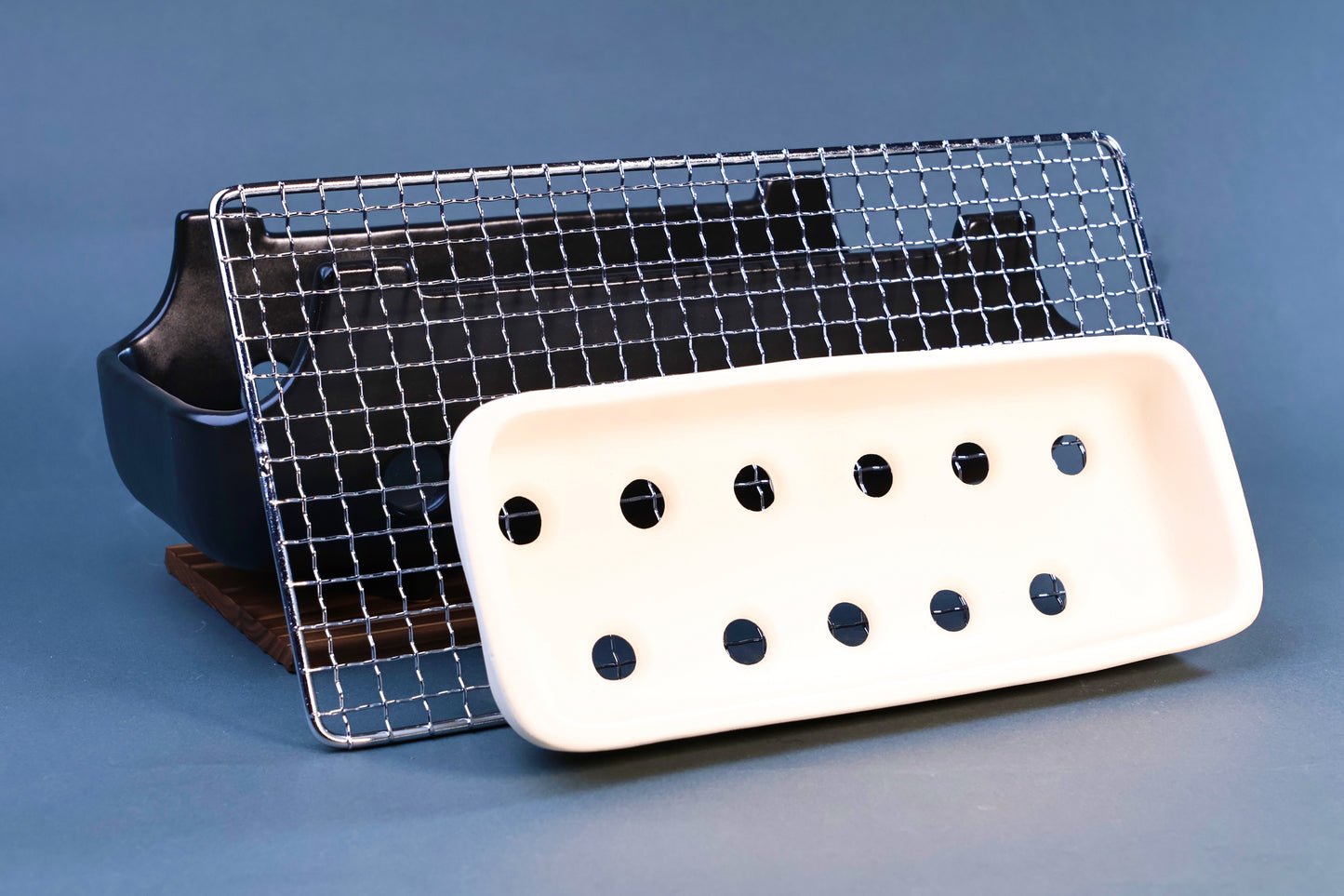 Banko yaki konro grill components showing black ceramic body, stainless wire grill and white ceramic fire plate on wood base against blue-grey background