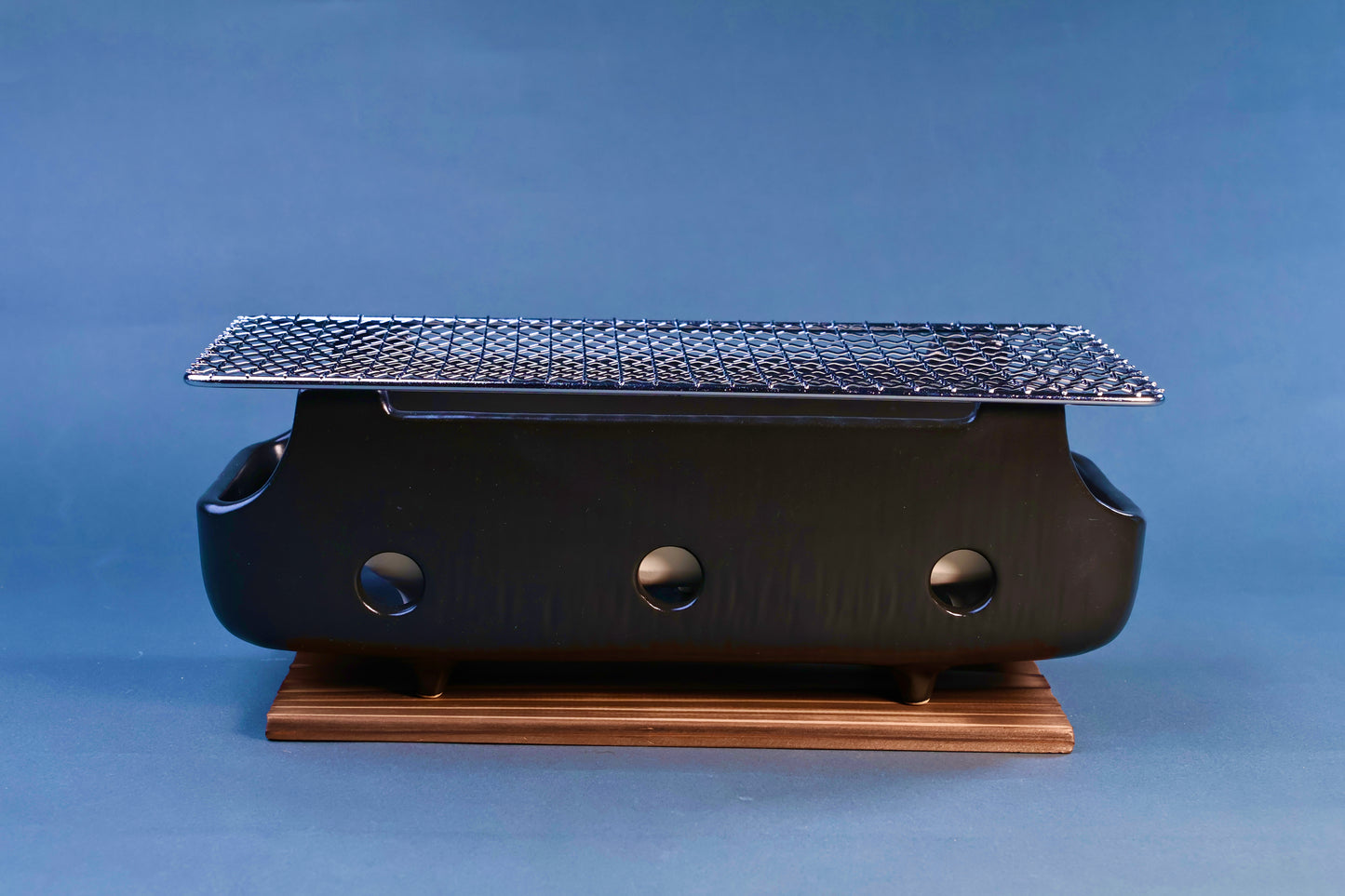 Banko yaki konro grill front profile showing black ceramic body with three ventilation holes, stainless wire grill and wood base on blue background