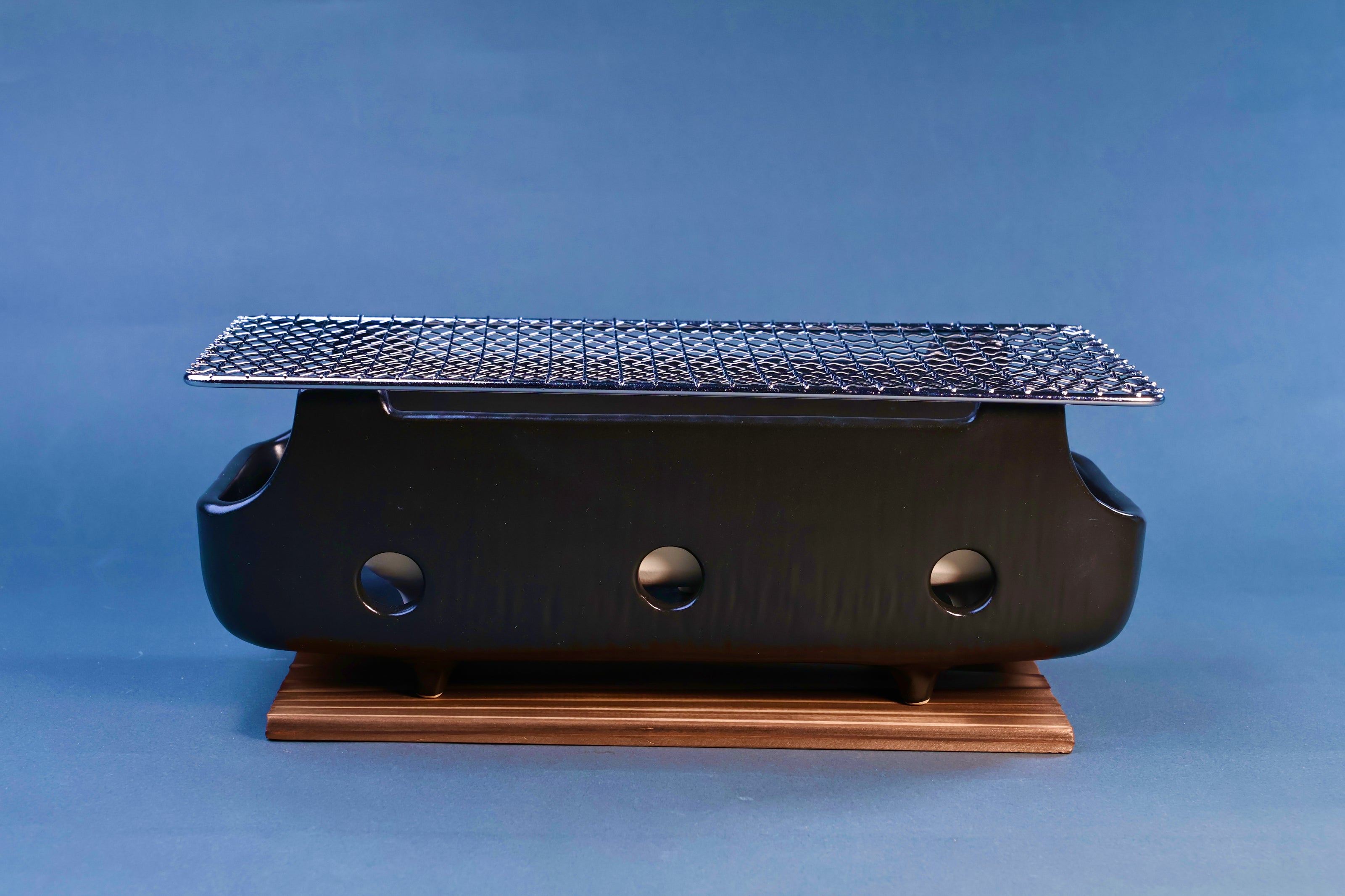 Banko yaki konro grill front profile showing black ceramic body with three ventilation holes, stainless wire grill and wood base on blue background