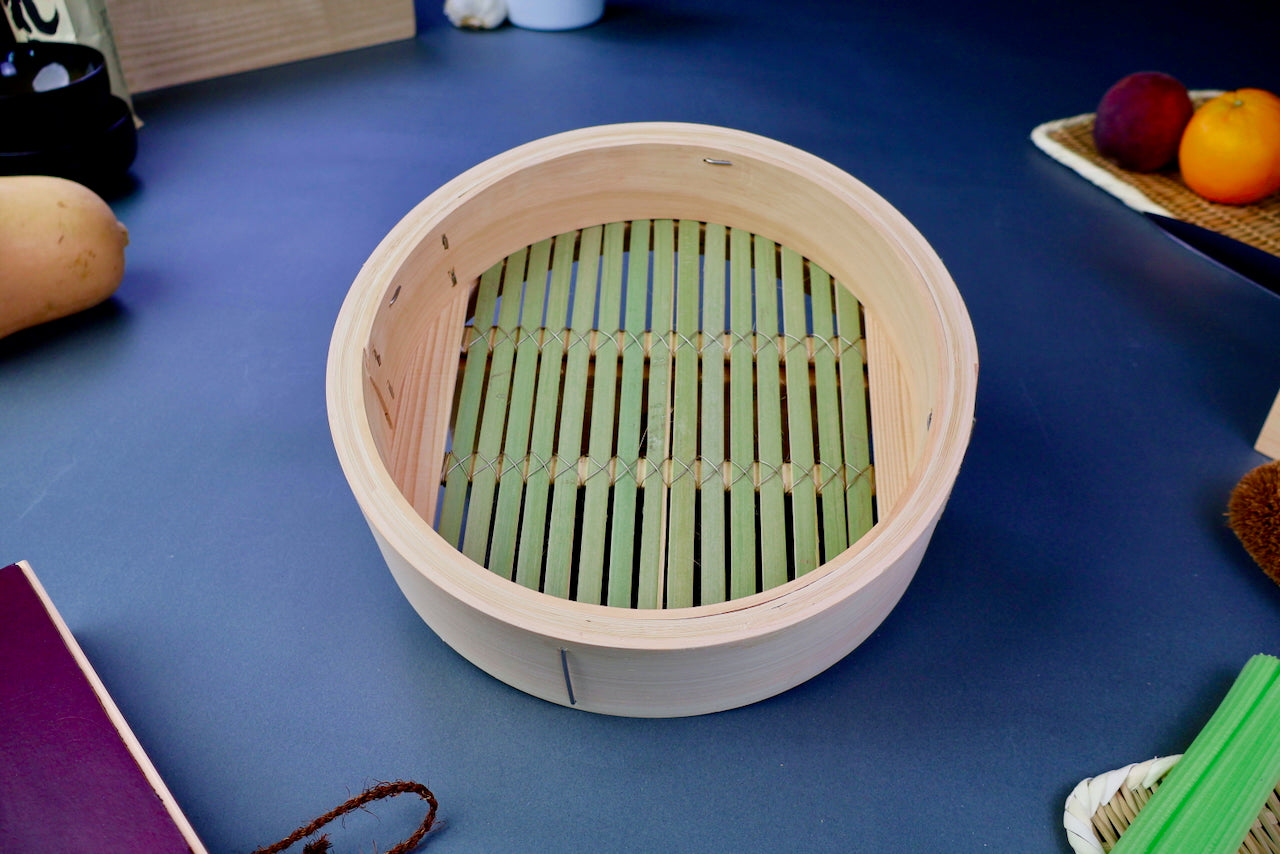 round thick hinoki wood bamboo steamer base with thick greenish slats interior surrounded by japanese kitchenware on blue gray surface